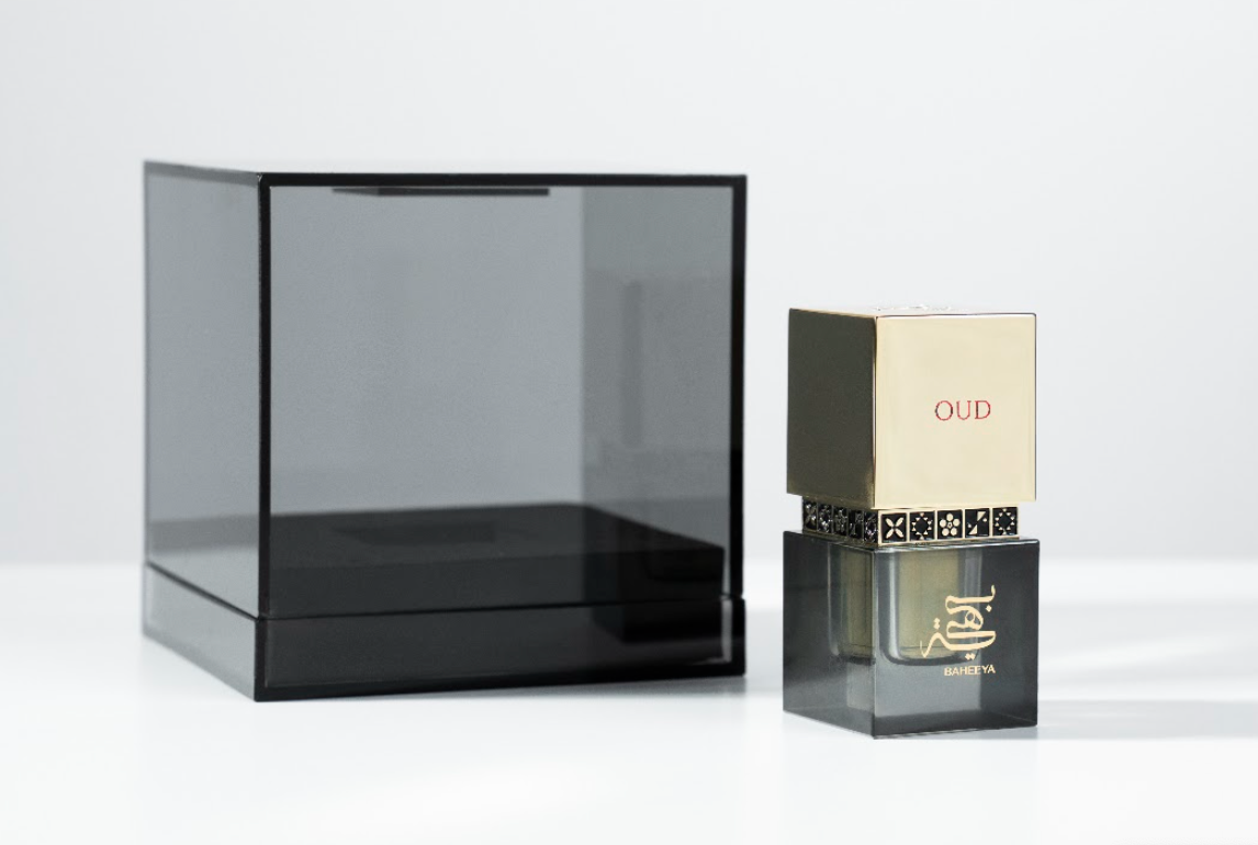 A perfume bottle with a gold cap and black patterned band next to a large transparent smoky black display box on a clean white background.